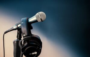 Ein Mikrofon, an dem ein Kopfhörer hängt vor verwaschenem, grau-blauem Hintergrund.