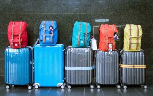 Fünf graue und blaue Rollkoffer in verschiedenen Größen stehen in einer Reihe an der Wand. Auf jedem Koffer befindet sich ein andersfarbiger Rucksack.