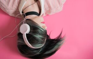 Eine Frau mit zerzausten Haaren liegt verkehrt herum im Bild: auf den Ohren trägt sie rosafarbene Kopfhörer.