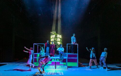 Eine Szene, in bläuliches Licht getaucht, aus dem Tanzstück „Time to Shine“ am Jungen Schauspiel Düsseldorf. Auf und um eine gerüstartige Bühne herum vollführen Männer und Frauen  unterschiedliche Bewegungen.